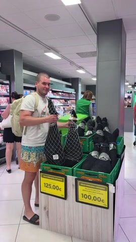 A man stands in a supermarket meat department holding two Spanish hams, one in each hand.  He is smiling and looking at the camera. He is wearing a white t-shirt, patterned shorts with images of melting ice cream, and black sandals. He has a beige backpack. He is standing in front of two green plastic crates filled with more hams encased in black netting. Price signs of 125,00€ and 100,00€ are visible. In the background, refrigerated shelves are stocked with packaged meats, and other shoppers are present. The lighting is bright, and the supermarket appears modern.