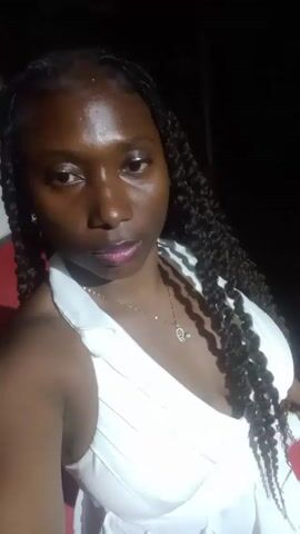 A woman with dark skin and long, dark braids is the subject of a selfie video. She wears a white, sleeveless dress and a gold necklace with a heart-shaped pendant. She is positioned against a dark background, and she raises her hand to her hair. The sound of music can be heard in the background.
