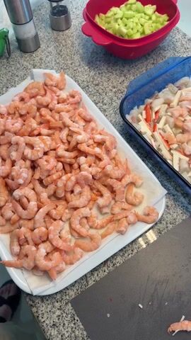 The video shows a kitchen counter with various ingredients and containers for preparing a seafood dish. A large white platter is filled with cooked shrimp. A blue glass dish contains cut-up imitation crab meat, shrimp, and a clear liquid, likely marinade or sauce. Another red container holds chopped cucumbers. A person's hand moves some of the shrimp on the platter. A knife and cutting board are also visible. The countertop is gray with a speckled pattern. The video shows the food items in close-up.
