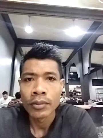 Here's a description of the video:

A man is filming himself in what appears to be a cafe or restaurant. He is wearing a black t-shirt and has short, dark hair. The background shows other patrons sitting at tables. The interior features archways, overhead lighting, and what looks like a black and white color scheme.  The camera moves slightly, showing more of the cafe interior at times.  The man appears to be looking directly at the camera while recording.