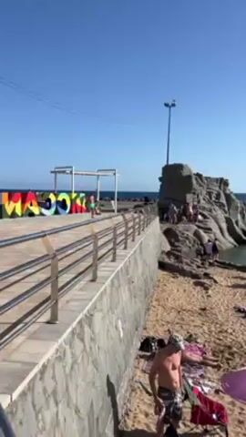 The video depicts a sunny day at a beach in Madeira. People can be seen sunbathing on the sand, swimming in the water, and playing in the sand near the shore. The beach is sandy with a few rocky formations along the coastline. There are a few structures that look like docks in the distance. In the first few moments of the video, you can see large block letters spelling out "Maco", the tail end of which is obstructed by a railing.