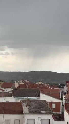 The video captures an overcast day over a town. A foreground view reveals the rooftops of closely packed houses, predominantly with red tile roofing. In the background, a ridge of hills stretches across the horizon, partially obscured by a hazy atmosphere. The sky is filled with thick, dark gray clouds, suggesting an impending storm. There are faint streaks of rain visible in the distance. A bird flies across the scene from left to right.