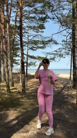 A woman stands in a forest and blows bubbles. She is wearing a pink tracksuit and white sneakers. She is holding a green wand. Behind her is a sandy path leading to a beach and the ocean. The sky is blue and the sun is shining. The video is shot at eye level and the camera is stationary.