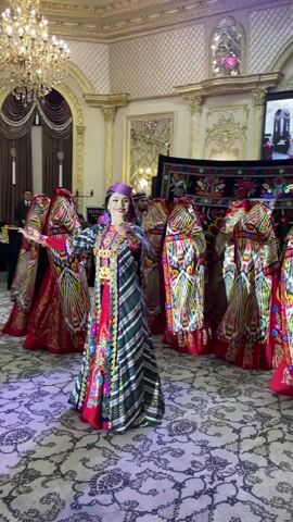 In a lavishly decorated room with ornate gold trim, chandeliers, and patterned carpets, a group of women in traditional, brightly colored ethnic attire perform a dance. The central dancer, in the foreground, wears a green, white, and black striped robe over a red dress, adorned with gold jewelry and a purple headscarf. She is actively engaged in dancing, her hands moving gracefully. Behind her, other dancers in similar vibrant red and patterned outfits, with covered heads, mirror her movements in a semi-circle. The background shows other individuals, some seated at tables, and decorative tapestries hanging on the wall. The lighting is theatrical, with purple light illuminating the dance floor.