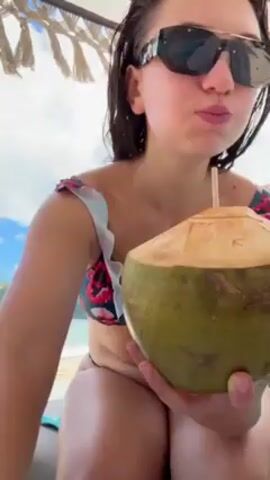 The video shows a woman in a floral bikini and sunglasses sitting on a beach chair under a thatched umbrella, drinking from a coconut. The beach and ocean are visible in the background.