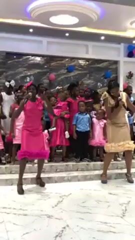 The video shows two women dancing and singing on a marble floor in a decorated room. Both women are holding microphones.  The woman on the left is wearing a bright pink dress and heels. The woman on the right is wearing a gold dress and blue shoes. Behind them, a group of children in pink dresses, a boy in a blue shirt, and adults are gathered on a tiered platform. The room is decorated with balloons. The ceiling has a modern circular light fixture.