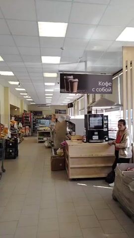 The video shows the interior of a store, likely a grocery or convenience store. The camera pans from right to left, revealing aisles stocked with products, checkout counters, and a coffee machine. A person is standing near the coffee machine, and another person is visible near a checkout counter in the background. The store appears to be mostly empty, with no other customers visible in the aisles. The lighting is bright, with fluorescent lights on the ceiling. Signs are visible in Russian, indicating product promotions and store information. The overall impression is of a quiet and uneventful shopping environment.