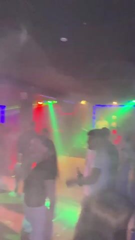 This is a blurry, low-light video of people dancing in a club. The atmosphere is dark with colorful flashing lights from above, including red, green, and blue beams. Some individuals are visible, but their faces and details are obscured by the blur and low lighting. The people appear to be dressed casually, and the focus is on the general ambiance of a lively party.