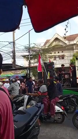 The video shows a street scene with a group of people gathered around a sound system mounted on a truck. A red and white Indonesian flag is prominently displayed on a pole next to the sound system. Several people are on motorcycles, some are standing around, and others are walking. The truck appears to be part of an event or celebration. The background shows buildings and utility poles. The overall atmosphere appears to be lively.