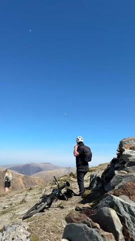 A mountain scene shot under a clear blue sky shows a man in a black outfit and a white helmet standing on a rocky slope, watching two paragliders in the sky. He's wearing a black backpack. A mountain bike lies on its side beside him. A woman stands some distance away on the same slope. The background features a hazy view of distant mountains. The ground is covered with rocks and sparse vegetation.