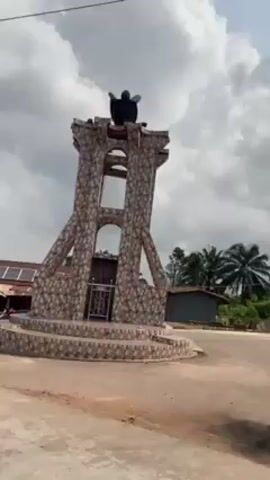 The video depicts a tall, stone-like monument with an open structure and a statue on top, situated at a road intersection in a rural area. The sky is overcast with heavy clouds. Palm trees and other vegetation are visible in the background, along with small buildings. A person is riding along the road and recording the scene. There are utility lines overhead. The road is paved and appears somewhat worn.