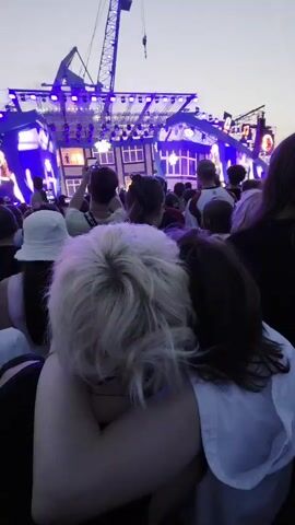 A first-person perspective shot from behind a crowd of people at an outdoor concert. The camera is looking towards a large stage with bright blue and purple lights. A crane is visible behind the stage structure. In the foreground, two people are embracing. The person closer to the camera has light blonde hair, and the person behind them has dark hair and is wearing a white top. The overall atmosphere is dark, with the stage lighting being the primary illumination. The crowd is densely packed, with many people holding up their phones.