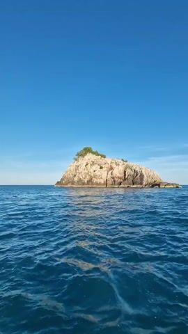 The video begins with a wide shot of a small, rocky island with sparse vegetation on top, surrounded by a vast expanse of blue ocean under a clear blue sky. The camera then pans to the right, showing more of the island's rocky coastline and continuing to move across the water. The camera continues to pan, revealing more distant landmasses on the horizon, also under a clear blue sky with a few wispy white clouds. The scene is one of a boat moving through calm, blue water, capturing the natural beauty of islands and the ocean.