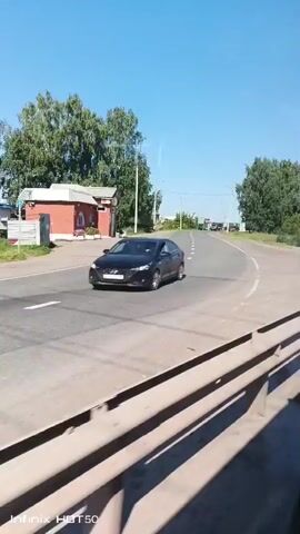 The video begins with a view of a paved road with white lane markings. A single-story building with a red roof and a small kiosk structure is visible on the left side of the road. Trees and vegetation line the sides of the road. The sky is clear and blue. As the video progresses, the camera appears to be moving forward along the road, possibly from inside a vehicle. The perspective shifts, showing more of the road and the surrounding greenery. A black car is briefly visible in the distance on the right side of the road. Streetlights are present along the roadside. The scene suggests a rural or suburban environment on a sunny day.
