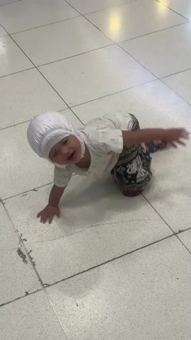 A child wearing a white cap and a patterned outfit is crawling on a tiled floor. The child is in motion, appearing to be falling or rolling. The floor is made of large, light-colored tiles with visible grout lines. The background is out of focus but appears to be an indoor space with overhead lighting.