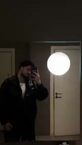 A man with brown curly hair and a beard is taking a mirror selfie. He is wearing a black leather jacket over a white t-shirt and dark pants. The lighting in the room is dim except for a bright, round light fixture on the wall to the right. There are two doors visible in the mirror, one to the left and one to the right, both white with silver handles. The wall is a neutral brown. The man is holding a phone with a dark green case and has rings and a bracelet on his hand. He smiles briefly.