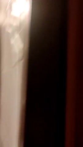 The video appears to be shot in a bathroom. The camera is pointed down towards the floor from a standing position. The bathroom features a white toilet, a patterned rug, and a pile of clothes. A pair of blue and white sneakers are visible on the rug.  The person filming is wearing a black hooded sweatshirt with white markings on the sleeve. The view is somewhat unsteady, suggesting the camera is hand-held.