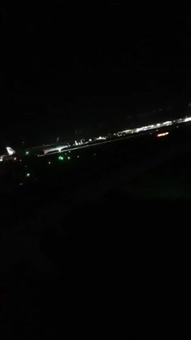At night, the blurred view from an airplane window shows an airport runway with various lights. The runway is lined with green and red lights. There are also white lights visible in the distance, possibly from buildings or other structures at the airport. The foreground is dark, and the scene is somewhat shaky. The video appears to be taken during taxiing or shortly before takeoff/landing, as the lights of the runway are visible but not in sharp focus.