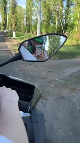 A first person view shows a person in a white t-shirt operating an all-terrain vehicle on a black asphalt road. The road has dirt and dead grass on both sides, then grassy areas and a line of tall trees. The person is operating the ATV from the left handlebar with their left hand and appears to be looking into the side mirror with their right hand holding up a cell phone. The mirror reflects the image of the driver in a helmet, a white t-shirt, and the cell phone being held up. The cell phone has a pink case with a cartoon bear on it. There is what appears to be animal dung or horse manure on the side of the road near the camera.