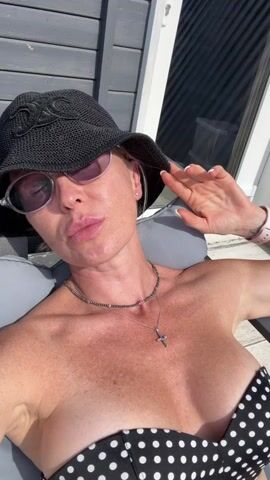 A woman wearing a black wide-brimmed hat, sunglasses, a beaded necklace, and a black and white polka-dot bikini top is lounging on a grey cushioned chair outdoors. She is looking at the camera and holding her right hand up to the brim of her hat. The background shows a grey wooden wall and a portion of a white door or window. Sunlight is casting shadows on the scene.