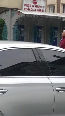 A silver car is parked in front of three public payphones. A woman wearing a red jacket and a headscarf stands by the payphones, looking to the right. A man wearing a blue jacket and a grey shirt is visible behind the car, also near the payphones. The payphones have graffiti on them. The sky is overcast.
