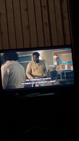 A television screen displays a scene from a movie or TV show in a dimly lit room. The screen illuminates two men in what appears to be an indoor setting, possibly an office or workshop. The man on the left is mostly seen from behind, with dark, curly hair and a light-colored, patterned shirt. The man on the right, with dark skin, a beard, and a light brown shirt, is facing slightly to the side. Behind him, a map of the United States is pinned to a wall, alongside other items on a desk or counter.

Bulgarian subtitles are visible on the screen. The first line reads: "Как намирането на кости" (How finding bones). The second line continues: "ни помага да се приберем у дома?" (helps us go home?). A third line of text is partially visible below, reading: "[Вс]е още не съм напълно сигурен." (Still not entirely sure.).

The background of the room where the television is located features a wall covered with striped wallpaper adorned with star-shaped decorations. The overall low light suggests the image was captured during the evening or night. A remote control is partially visible at the bottom, just below the television screen.