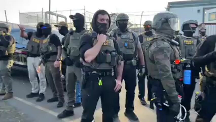 A group of approximately a dozen law enforcement personnel, possibly from Immigration and Customs Enforcement (ICE) and its Enforcement and Removal Operations (ERO) division, stands in an outdoor area. Many of the individuals are wearing tactical vests with "POLICE ICE" and "ERO" patches, along with helmets, balaclavas, and other protective gear, obscuring their faces. They are equipped with various items attached to their vests, including what appear to be radios, spray canisters, and other tools.

The setting appears to be a paved area, possibly a street or parking lot, adjacent to a chain-link fence and a building. Bright sunlight, possibly from a setting sun, casts a strong glare and creates long shadows, suggesting late afternoon or early evening. A vehicle is partially visible in the background on the left. The officers are generally facing forward, with some looking towards the sides, and one individual in the foreground is seen adjusting their gear. Chants of "All power to the people" and "They have proven they want to hunt and cage and destroy themselves" are audible, indicating a protest or demonstration occurring in the vicinity.