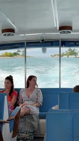 The video shows the interior of a boat moving through water. The boat has blue seats and a white ceiling with circular vents. There are three women sitting on the seats. The woman in the middle is wearing a floral dress and has a watch on her left wrist. The woman to her left is wearing a bright orange top and has her hair up in a bun. Through the window behind them, there is a view of the ocean with small islands in the distance. The boat is creating a wake as it moves through the water. A security camera is mounted on the ceiling of the boat.
