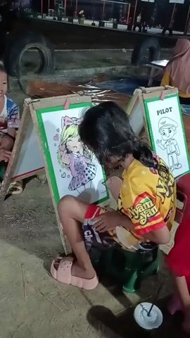 A young girl, wearing a yellow shirt and striped shorts, sits on a green stool and colors in a picture of an anime character. She is right-handed and is using a pen to color the drawing. In front of her is an easel with the drawing. To her left, another child, a boy, sits on a green stool and watches her. He is wearing a blue and white shirt. Behind them, there is a dark background with what appears to be a tire and some tarp.
