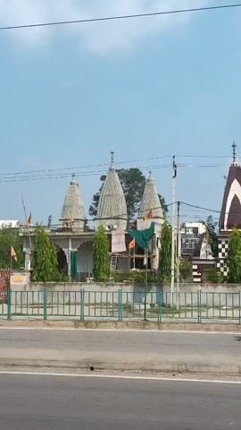 The video captures a street view of a temple with multiple spires. The temple appears to be made of stone with some surrounding trees and foliage. A red and white truck drives past the temple on the right side of the frame. A man on a motorcycle is visible in the distance on the road. The sky is mostly clear with some clouds.