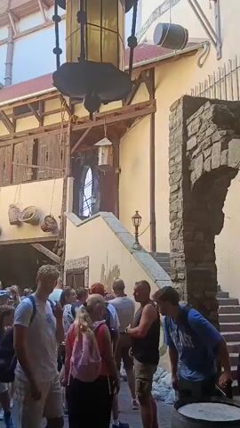 The video shows a courtyard with a group of people standing in line. The courtyard has stone walls, wooden structures, and a tower. There is a large hanging lantern in the center of the courtyard. The people in line are dressed in casual clothing, and many are carrying backpacks. The atmosphere appears to be that of an amusement park or historical attraction. The video pans slightly to the left and right, showing different parts of the courtyard and the people in line.