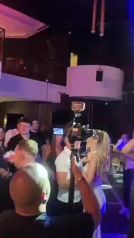 In a dimly lit indoor space, likely a nightclub or event venue, a group of people are gathered. A woman, dressed in a light-colored dress, appears to be the center of attention, dancing with someone whose face is partially obscured. Several people are watching and clapping. A person is filming the scene with a camera or phone. The venue has a raised platform, lighting fixtures, and architectural details that suggest a modern or luxurious setting. The color palette is dominated by blues and purples from the lighting, with darker tones throughout the rest of the venue. The atmosphere appears celebratory and social.