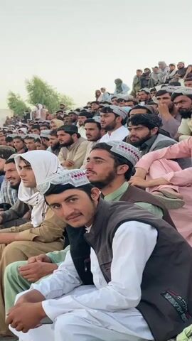 A crowd of men and boys are seated in rows, appearing to be spectators at an event. They are mostly wearing traditional clothing, including turbans, caps, and vests. The background consists of a neutral colored wall and a pale sky, with some trees visible on the left side. The focus is on the faces of the individuals in the foreground, many of whom are looking towards the camera. The overall impression is of a gathering or public assembly.