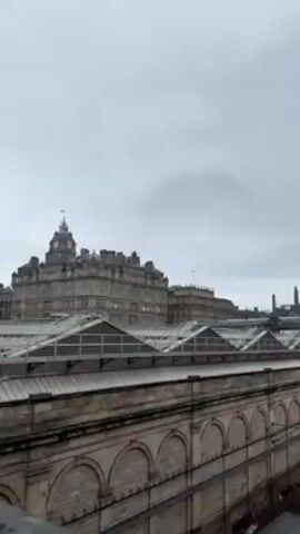 The video shows a view of a train station from an elevated position. The camera slowly pans to the right, revealing more of the station and the cityscape in the background.  The station platforms appear wet, suggesting it has recently rained.  The sky is overcast and grey.  In the background, a tall building with a clock tower and other buildings can be seen.  There are no people visible in the video.  The architectural style of the station and surrounding buildings suggests a European city.