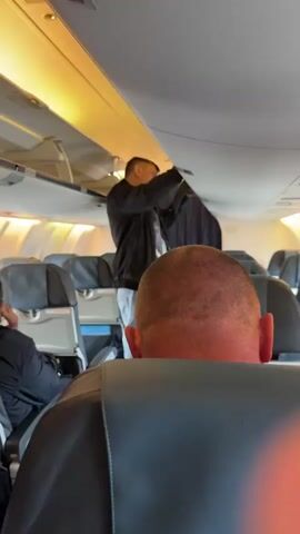 A man in a black jacket and white pants is standing in the aisle of an airplane cabin, pulling down a dark garment from the overhead compartment. Rows of empty passenger seats with dark headrests are visible. The overhead bins are open, revealing luggage. The lighting is warm and yellowish. The camera is positioned from the perspective of a seated passenger, looking over the headrest of the seat in front.