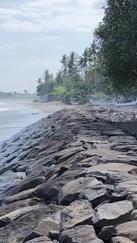 The video shows a wide shot of a rocky coastline meeting the ocean under a bright, slightly cloudy sky. In the foreground, a slope of large, grey rocks forms a barrier against the water. Waves crash against these rocks and then recede. To the left, the ocean extends out with more waves visible. To the right, beyond the rocky barrier, a sandy beach is visible, backed by a line of palm trees and other green foliage. Several small, light-colored fishing boats are pulled up on the sand near the treeline. The scene is calm with the continuous motion of the waves.