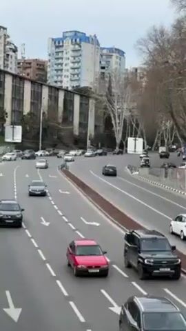 The video depicts a busy multi-lane highway in a city setting. Cars, including sedans and SUVs of various colors (red, white, black, gray), are actively moving along the road, following the painted lane markings and directional arrows. A white minibus also travels among the cars. The background shows apartment buildings of different heights and architectural styles, along with trees (some bare) lining the roadside. The sky is overcast, and the overall atmosphere seems urban and functional. The shot is taken from an elevated position, giving a broad view of the traffic flow and the surrounding cityscape.