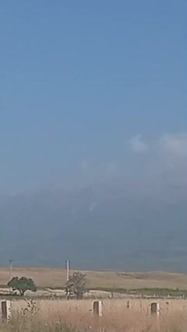 The video is a landscape shot of a dusty, dry field with tall, dead grass in the foreground. In the background, there are rolling hills and distant mountains, shrouded in haze. The sky is a clear, pale blue with a few scattered white clouds. There are a few man-made structures visible, including what appear to be fence posts or small concrete pillars in the foreground, and a tall electrical pylon on the right side of the frame. The overall impression is one of a vast, arid, and undeveloped area.