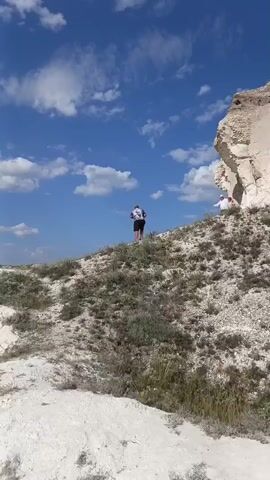 The video shows a rocky, arid landscape under a bright blue sky with scattered white clouds. Two people are visible on a grassy, white-rock slope. The person in the foreground, wearing a dark shirt with a white pattern and dark shorts, stands facing away from the camera, appearing to hold a phone or camera. Further up the slope, another person in a white shirt and dark pants is seated. The terrain is uneven, with sparse vegetation and a winding white path leading upwards. The camera pans across the landscape, focusing on the white rock formations and the surrounding hills.