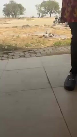 The video shows an outdoor scene with a person standing on a tiled porch or patio. The person is wearing black pants and black shoes with a silver buckle. The person is also wearing a colorful top with a flame-like pattern, although only a portion of it is visible. Outside the porch is a dry, grassy area with scattered debris and two trees in the distance. A motorcycle is parked in the grassy area. The ground is covered in a mix of dirt and dry grass. The scene is shot from a slightly low angle.
