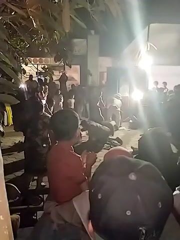 A group of people are gathered outdoors at night. The scene appears to be a courtyard or open area. There are several individuals in the foreground, some sitting and others standing. In the background, a larger crowd is visible, with many people actively engaged in what appears to be a heated or chaotic situation. Some individuals are in motion, possibly struggling or being held. Lights, possibly from a vehicle or lighting equipment, are illuminating the scene, creating bright glare and casting shadows. Trees and foliage are present in the upper portion of the frame, suggesting an outdoor environment. Clothing and hairstyles are diverse, indicating a mixed group of people. The overall atmosphere suggests a public gathering that has escalated into a disturbance.
