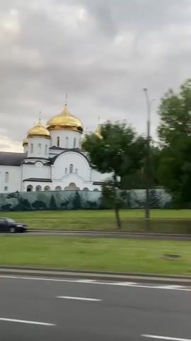 The video captures a perspective from a moving vehicle, likely a car, on a cloudy day. The primary subject is a large white church with several golden domes. The church features arched windows and a prominent facade. In front of the church, a dark green fence with a repeating pattern of trees and wildlife is visible.

To the left of the church, a tall, slender white pole, possibly a lamppost or utility pole, is the closest object to the camera and is blurred due to the motion. A dark-colored sedan drives along the road in front of the church. Further to the left, a tree with lush green foliage is also blurred by the movement.

The road in the foreground is asphalt with white lane markings. The scene suggests a suburban or semi-rural setting in Belarus, given the location context provided. The sky is overcast, indicating a cloudy or possibly overcast day. There are no people prominently visible in the frame. The overall mood is one of passing by a significant landmark, with the motion of the vehicle creating a dynamic visual experience.