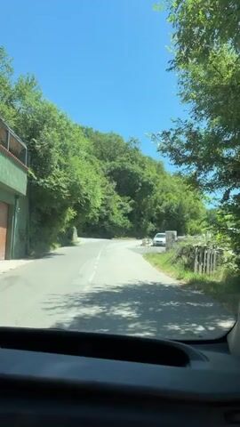 The video shows a view from inside a vehicle, looking out through the windshield. The vehicle is driving on a paved road that curves to the left, surrounded by lush green trees and foliage on either side. The sky is a clear, bright blue. A white car is parked on the right side of the road near a stone wall. Further ahead, another white car is visible driving away from the viewer on the same road. The road has white lane markings. There is a metal guardrail along the right side of the road. The interior of the vehicle is partially visible at the bottom of the frame, including the dashboard. The sun is shining brightly.