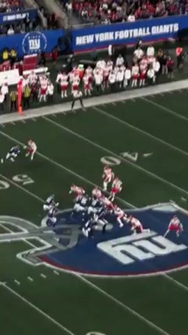 The video shows an aerial view of an American football game on a green field with white yard lines and a large team logo in the center. Players from two teams, one in red and white uniforms and the other in blue and white uniforms, are engaged in a play. Players are running and tackling each other. In the background, spectators are visible in the stands. A sign reads "NEW YORK FOOTBALL GIANTS". A referee is standing on the field.