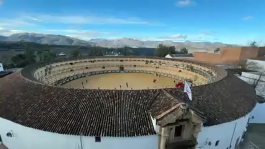 The video shows an aerial view of a bullring under a partly cloudy blue sky. The bullring is circular with a tan sand-covered arena surrounded by tiered seating. A few people are visible within the arena. The structure has a terracotta tiled roof with a small ornate building attached. In the background, a mountainous landscape is visible.
