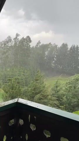 The video shows a view from a balcony during a heavy rainstorm or hailstorm. The sky is overcast and grey. Trees in the distance are swaying violently in the wind. The ground is obscured by the downpour. The balcony's railing is visible in the foreground, offering a glimpse of decorative cutouts.  The overall atmosphere is one of turbulent weather.