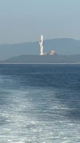 A very tall, white statue of a person stands prominently on a hill overlooking a body of blue water. To the right of the statue, there is a building with a curved, orange roof. In the distance, mountains form the backdrop against a clear sky. The water in the foreground is choppy, with white foam trails suggesting movement, possibly from a boat. Two small boats are visible in the distance on the water. The overall scene appears to be a coastal landscape on a bright, clear day.