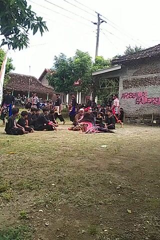 Here's a description of the video:

The video shows a group of people gathered in an outdoor area, likely a courtyard or field, with buildings visible in the background. Many people are sitting on the ground, while a smaller group is standing and appears to be engaged in some sort of performance or ceremony. 

The standing individuals are wearing distinctive black outfits with contrasting patterns, possibly traditional attire. Some of them are holding flags or banners. The surrounding environment includes trees, grass, and buildings with tiled roofs. The overall atmosphere seems communal and possibly celebratory.