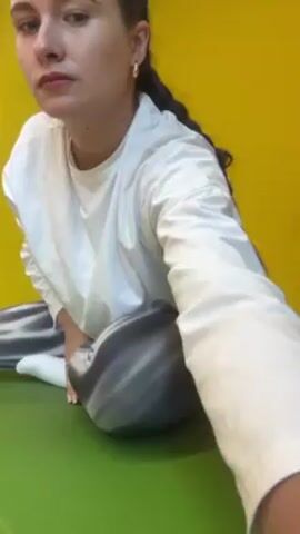 A woman is sitting on a green yoga mat in front of a yellow wall. She is wearing a white long-sleeved shirt and gray tie-dye patterned leggings. She leans to her right side, bending her torso sideways while keeping her left hand on her left thigh. Her hair is pulled back in a ponytail, and she has gold hoop earrings. A pair of white sneakers sits on the floor to the right of the mat.