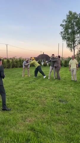 The video shows a group of men playing what seems to be a humorous game on a grassy lawn. There are five men standing around and watching. The first of the men is in a dark suit and looks to be filming. The second man is wearing a cowboy hat and a light yellow shirt and beige pants. The third man in the middle of the group is being lifted up by his legs as he hugs another man. The fourth man is hugging the fifth man in the group. There is a house in the background, as well as some trees. The sky is a light blue with a hint of pink and orange.